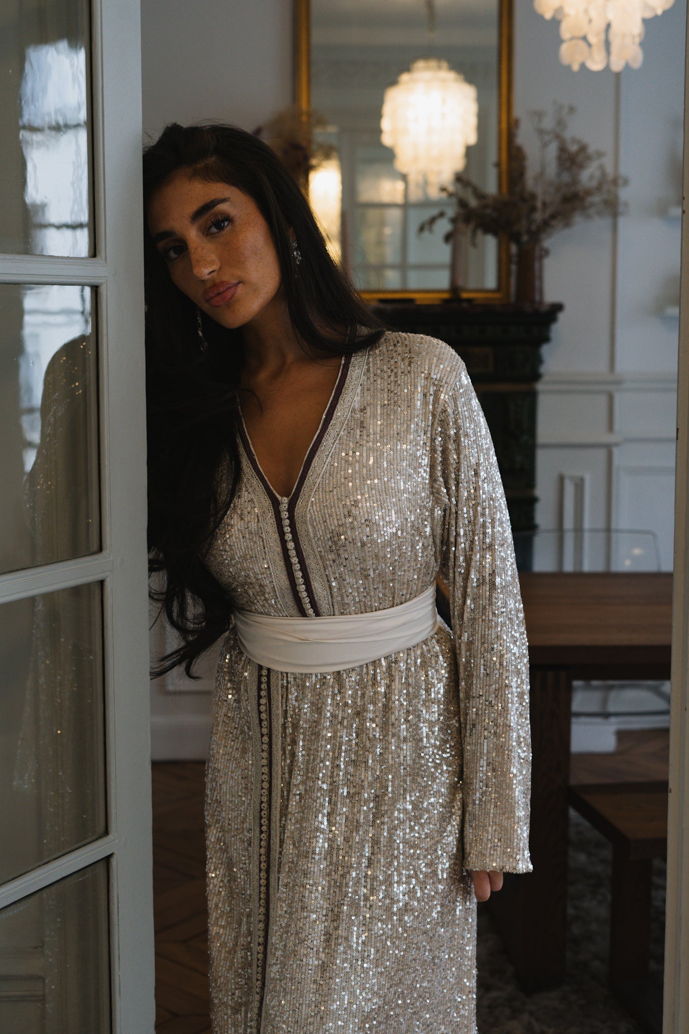 Une femme est adossée contre une porte, elle porte un caftan gris à sequin
