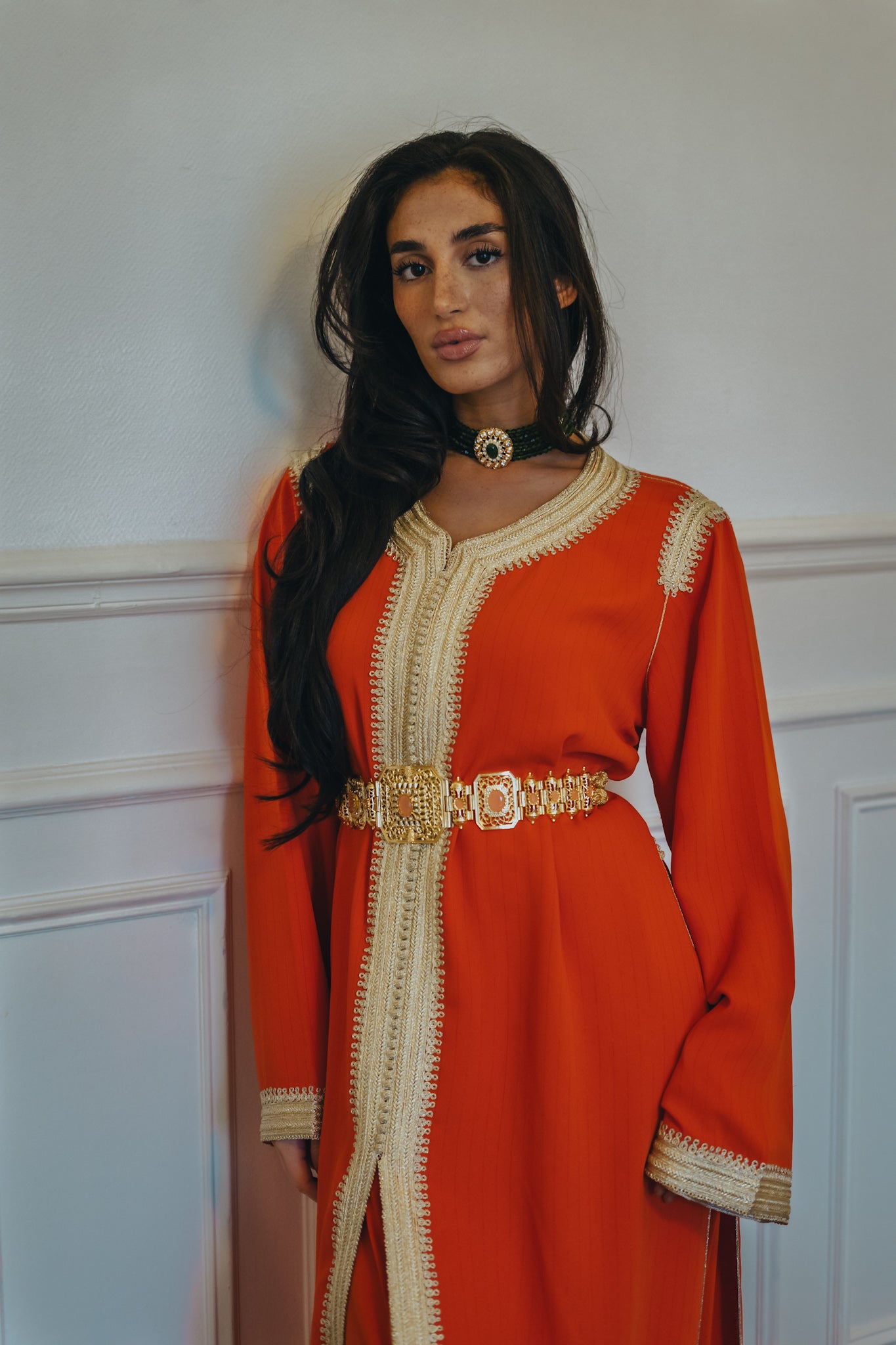 Une femme porte un caftan orange, un collier vert et une ceinture marocaine dorée
